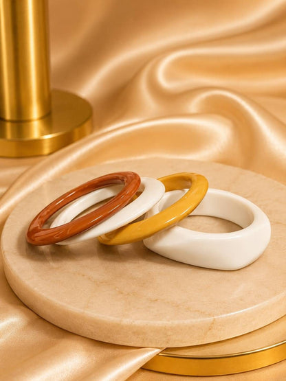 Three resin bangles in terracotta, golden amber and white fanned out on a marble disc with a gold satin background