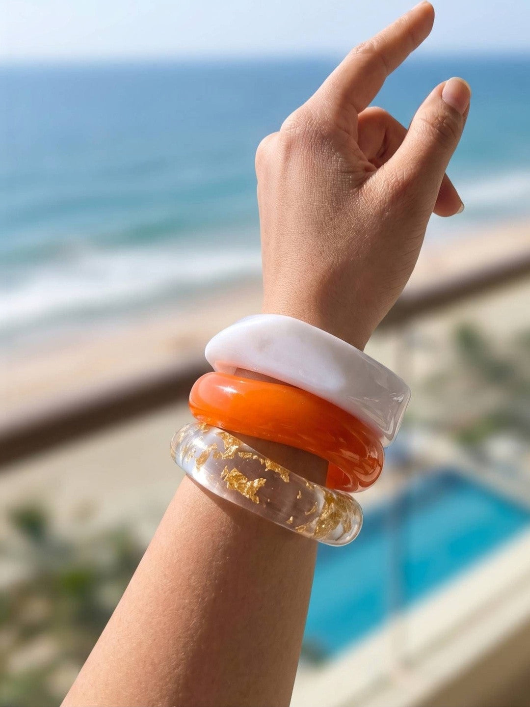 Three stacked resin bangles on a raised wrist against a beachside backdrop with ocean and pool visible
