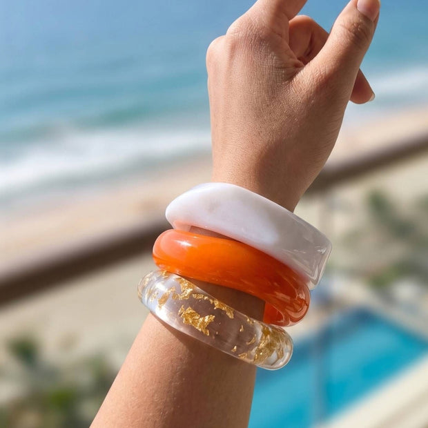 Three stacked resin bangles on a raised wrist against a beachside backdrop with ocean and pool visible