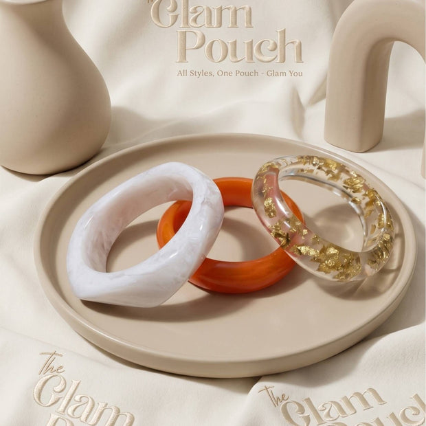 White marble, orange, and clear gold-foil resin bangles arranged on a beige tray with branded pouches