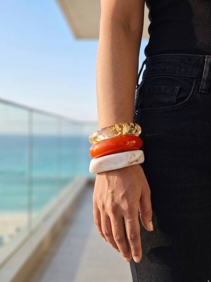 Three chunky resin bangles stacked on a wrist wearing black jeans with sea view in background