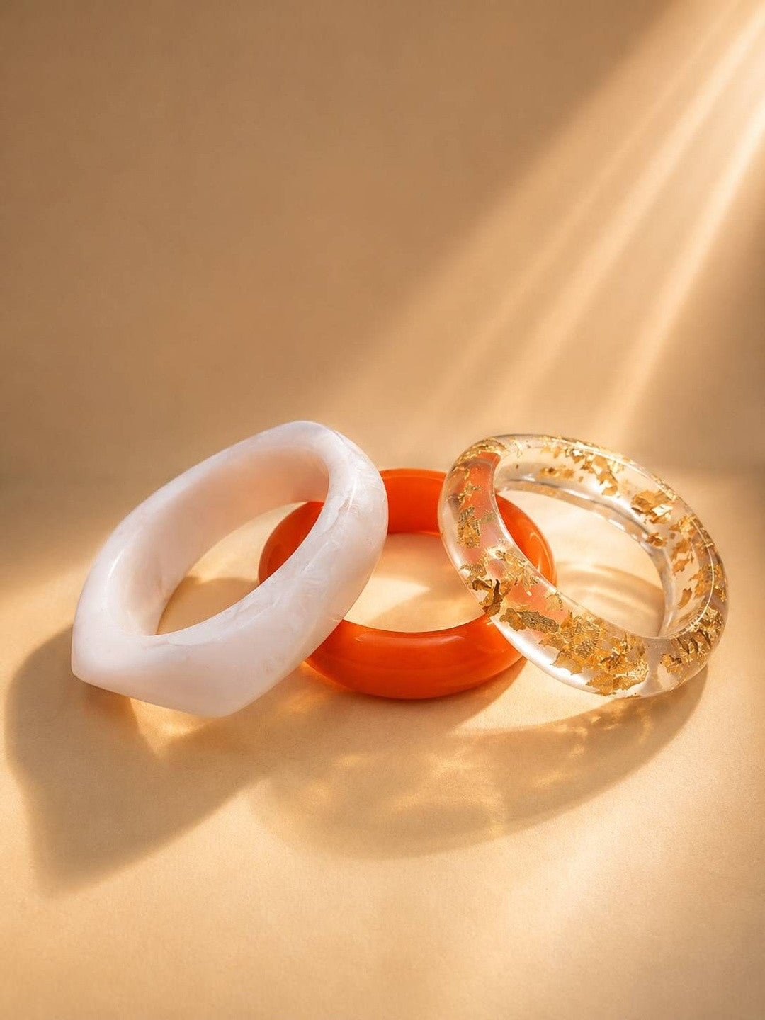 White marble, orange, and clear gold-foil resin bangles leaning against each other in warm directional light