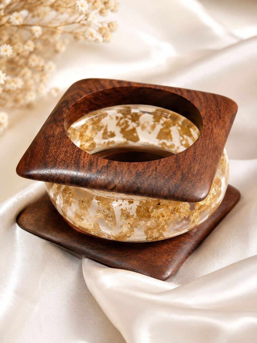 Clear resin bangle with gold foil flakes resting inside a square walnut wood kada on white satin fabric
