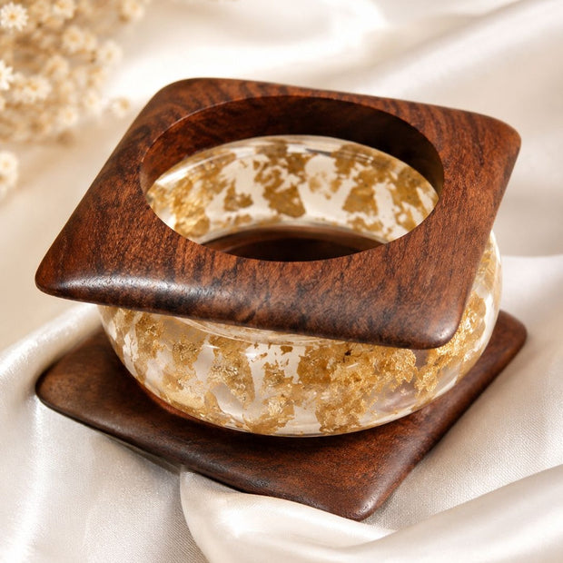 Clear resin bangle with gold foil flakes resting inside a square walnut wood kada on white satin fabric
