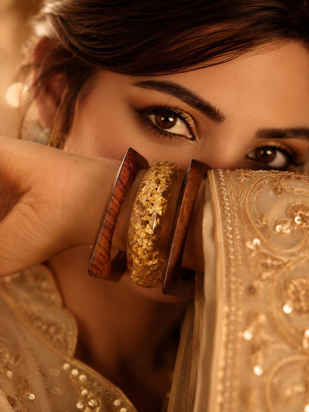 Close-up of woman in embroidered ivory lehenga holding gold foil resin and wood bangles near her face