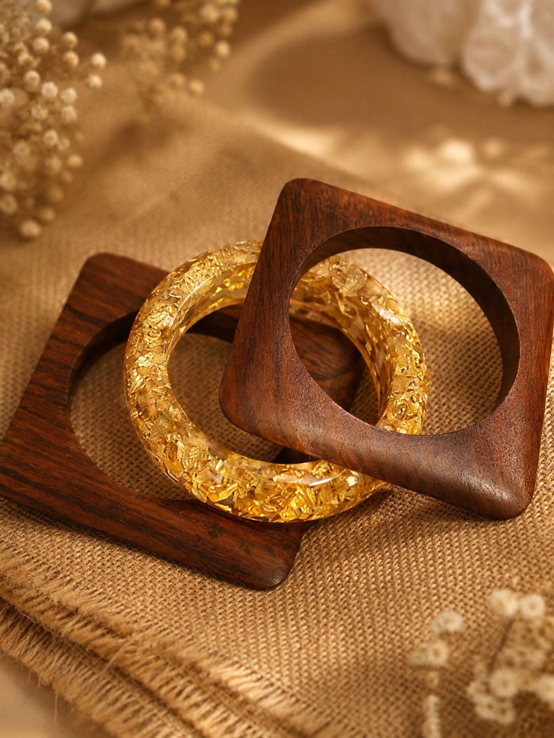 Two square wooden kadas and one round gold foil resin bangle laid on a wooden tray with dried flowers