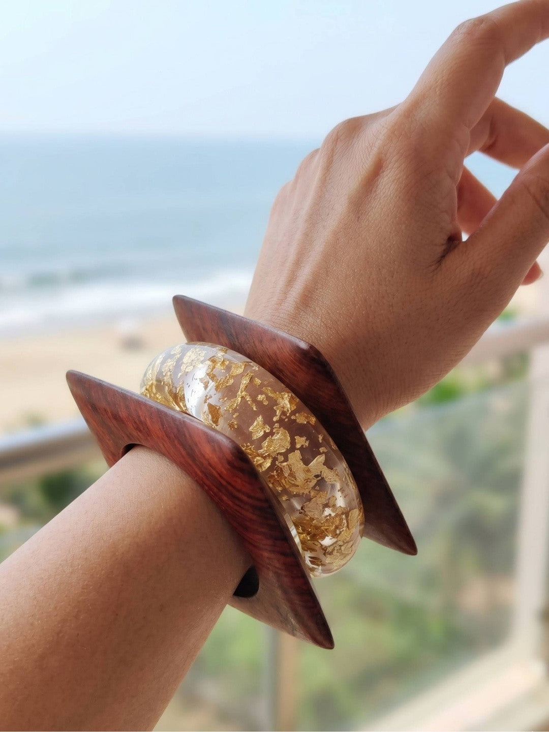 Gold foil resin bangle and square wood kada worn together on a wrist with a beach backdrop