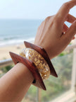 Gold foil resin bangle and square wood kada worn together on a wrist with a beach backdrop