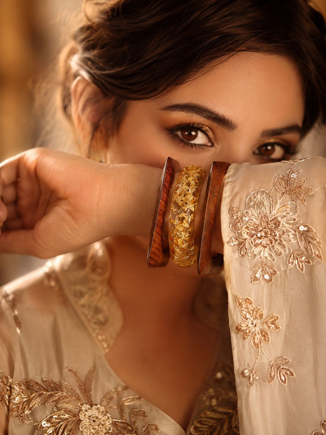 Woman in cream embroidered outfit holding gold foil resin and wood bangles stacked on her wrist near her face