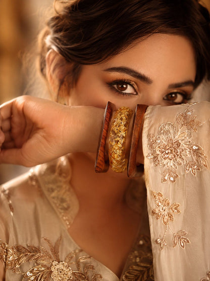 Woman in cream embroidered outfit holding gold foil resin and wood bangles stacked on her wrist near her face