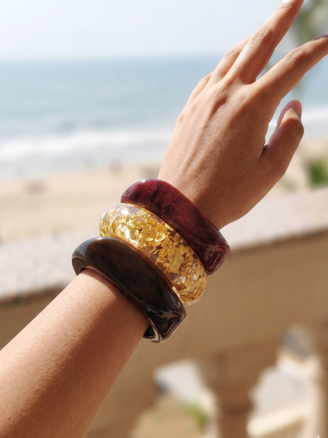 Three stacked resin bangles on a raised wrist against a beach backdrop, showing burgundy marble, clear gold-foil, and dark tortoise finishes