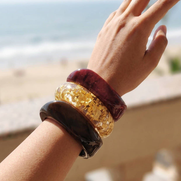 Three stacked resin bangles on a raised wrist against a beach backdrop, showing burgundy marble, clear gold-foil, and dark tortoise finishes