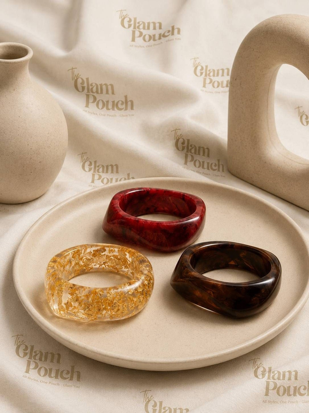 Flat lay of three resin bangles on a ceramic plate — burgundy marble, clear gold-foil, and dark tortoise — styled with neutral ceramic props