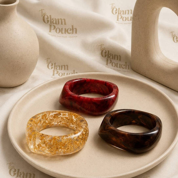 Flat lay of three resin bangles on a ceramic plate — burgundy marble, clear gold-foil, and dark tortoise — styled with neutral ceramic props