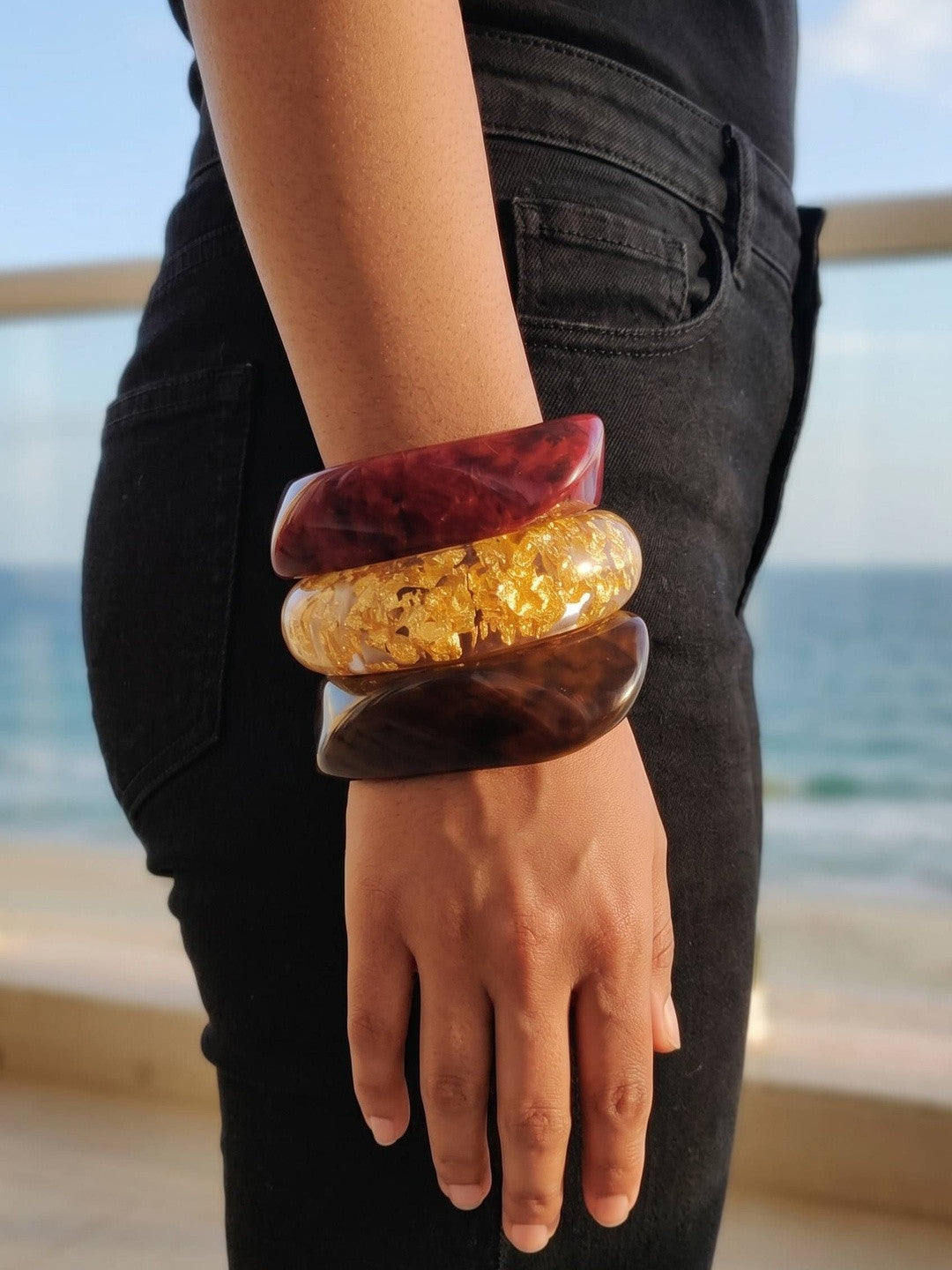Side view of three resin bangles stacked on a wrist against a black outfit and ocean background, showing chunky silhouette and high-gloss finish