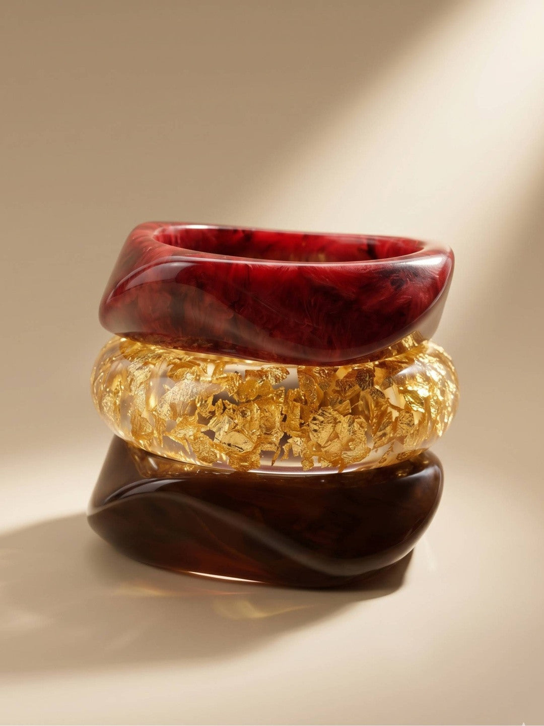 Close-up studio stack of three resin bangles — burgundy marble on top, clear gold-foil in the centre, dark tortoise at the base — under warm directional light