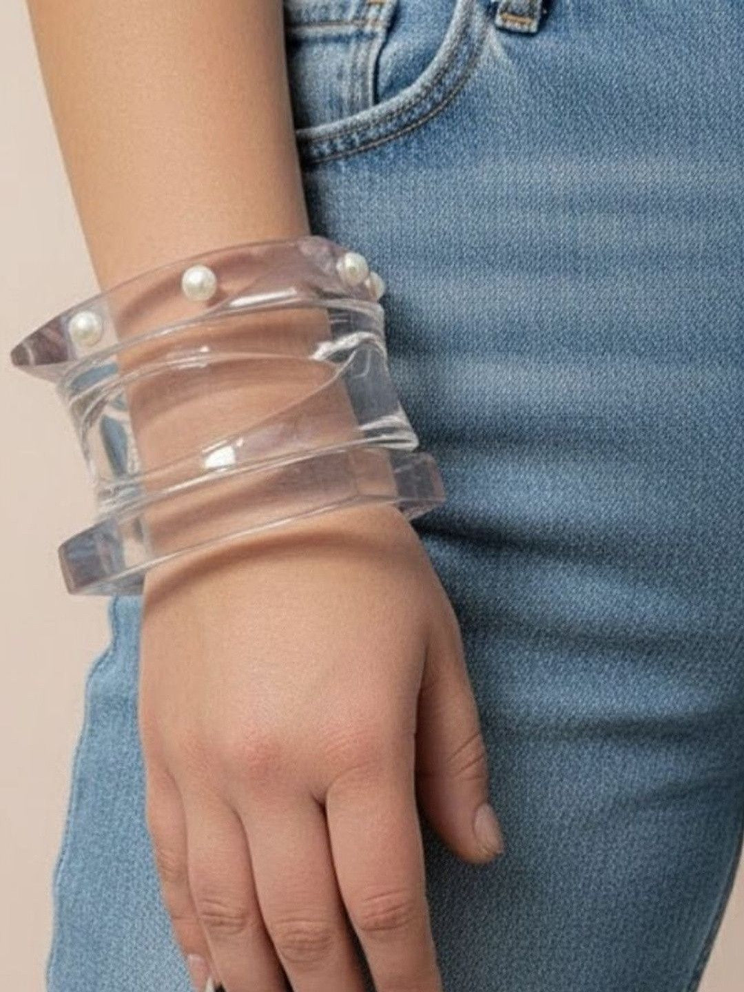 Three clear acrylic pearl stud bangles stacked on a woman's wrist against blue denim jeans