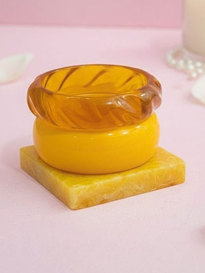 Three resin bangles in amber, mustard and marbled yellow stacked on a square yellow resin prop against a pink background
