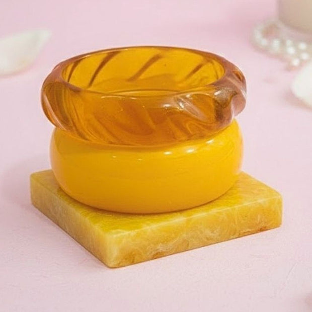 Three resin bangles in amber, mustard and marbled yellow stacked on a square yellow resin prop against a pink background