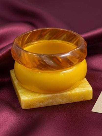 Close-up of translucent amber carved bangle resting on opaque mustard bangle on a yellow marble block against burgundy silk