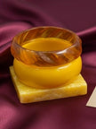 Close-up of translucent amber carved bangle resting on opaque mustard bangle on a yellow marble block against burgundy silk