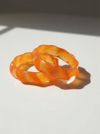 Two orange marbled resin bangles with wavy sculpted edges resting on a white surface in sunlight