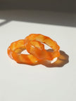 Two orange marbled resin bangles with wavy sculpted edges resting on a white surface in sunlight