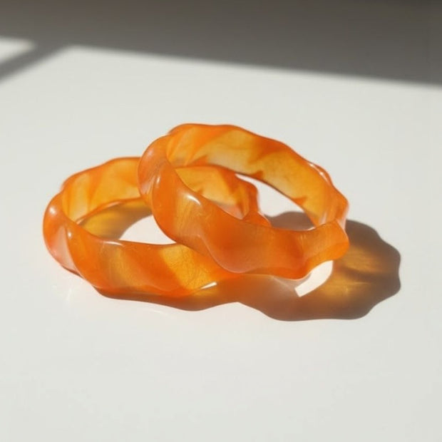 Two orange marbled resin bangles with wavy sculpted edges resting on a white surface in sunlight