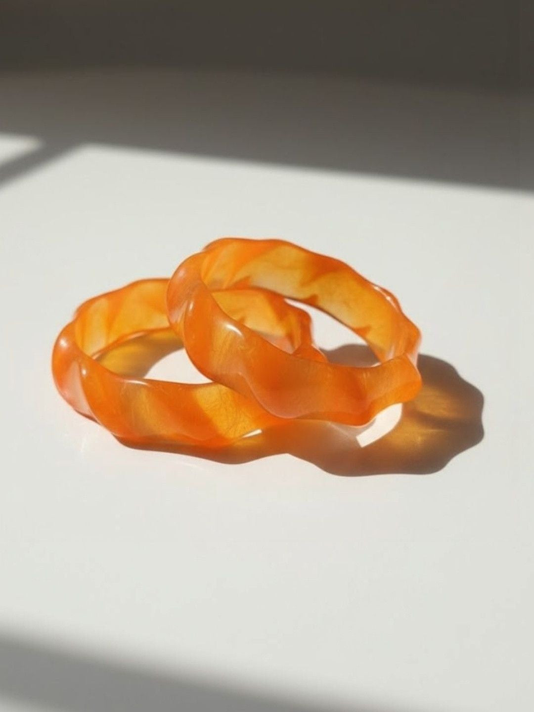 Two orange resin wavy bangles stacked together on a white surface with soft shadows