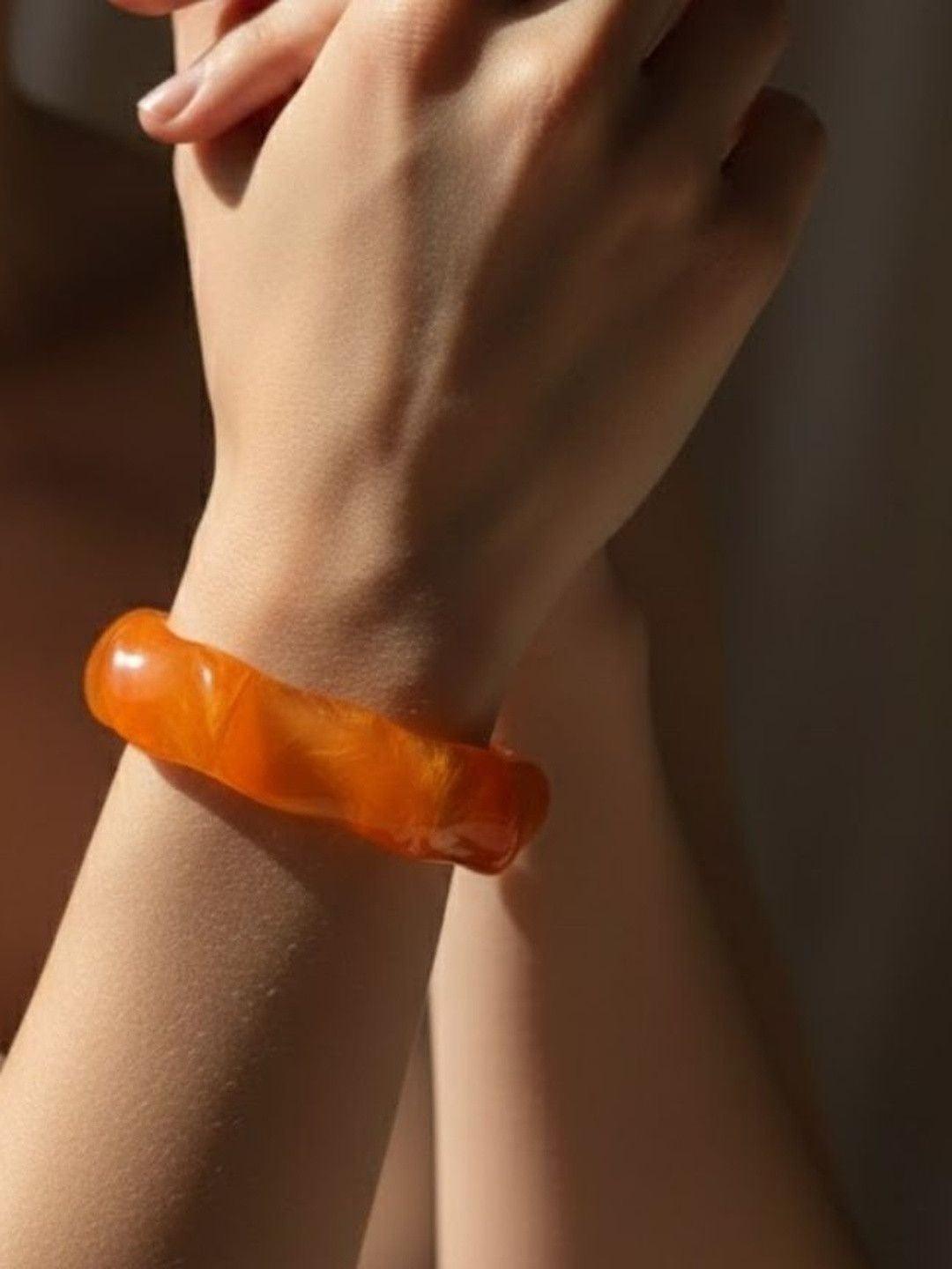 Orange resin wavy bangle worn on a woman's wrist with hands crossed against a dark background