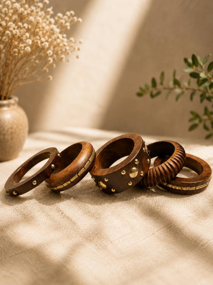 Five dark wooden bangles laid flat on linen fabric showing individual designs including studs, tiles, carvings and rivets