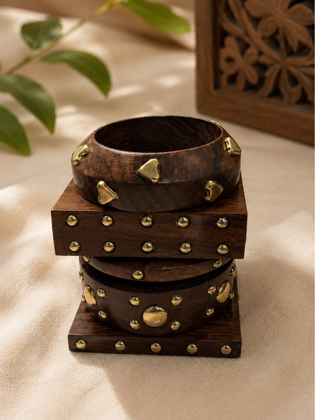 Three brass-studded wooden bangles stacked vertically near carved wooden decor and green foliage