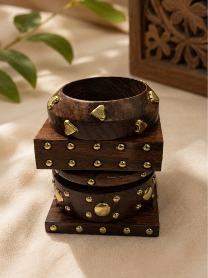 Three brass-studded wooden bangles stacked vertically near carved wooden decor and green foliage