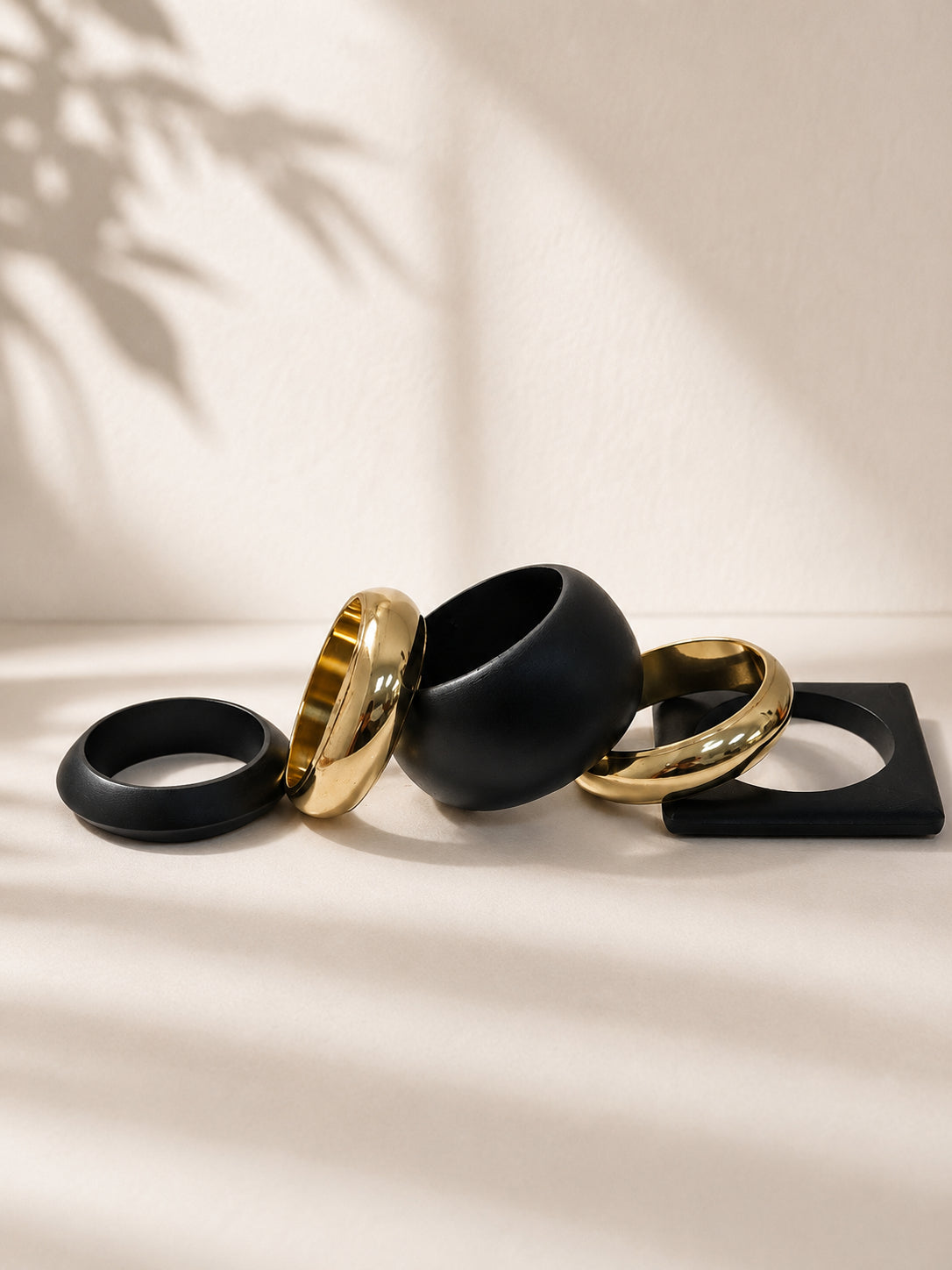 Five matte black and gold-tone bangles of varying shapes arranged flat on a cream surface with leaf shadow