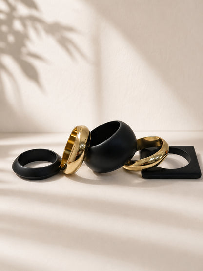 Five matte black and gold-tone bangles of varying shapes arranged flat on a cream surface with leaf shadow