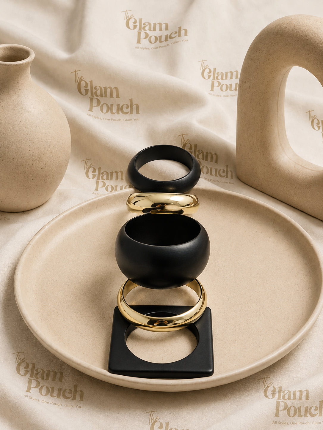 Five matte black and gold-tone bangles stacked vertically on a beige ceramic plate with The Glam Pouch branded backdrop