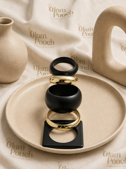 Five matte black and gold-tone bangles stacked vertically on a beige ceramic plate with The Glam Pouch branded backdrop