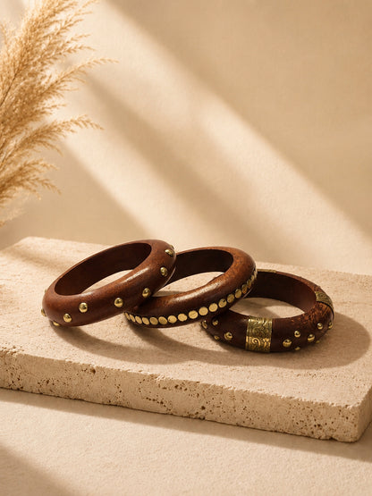 Three wooden bangles with brass studs and coin accents laid on a travertine surface with dried pampas grass