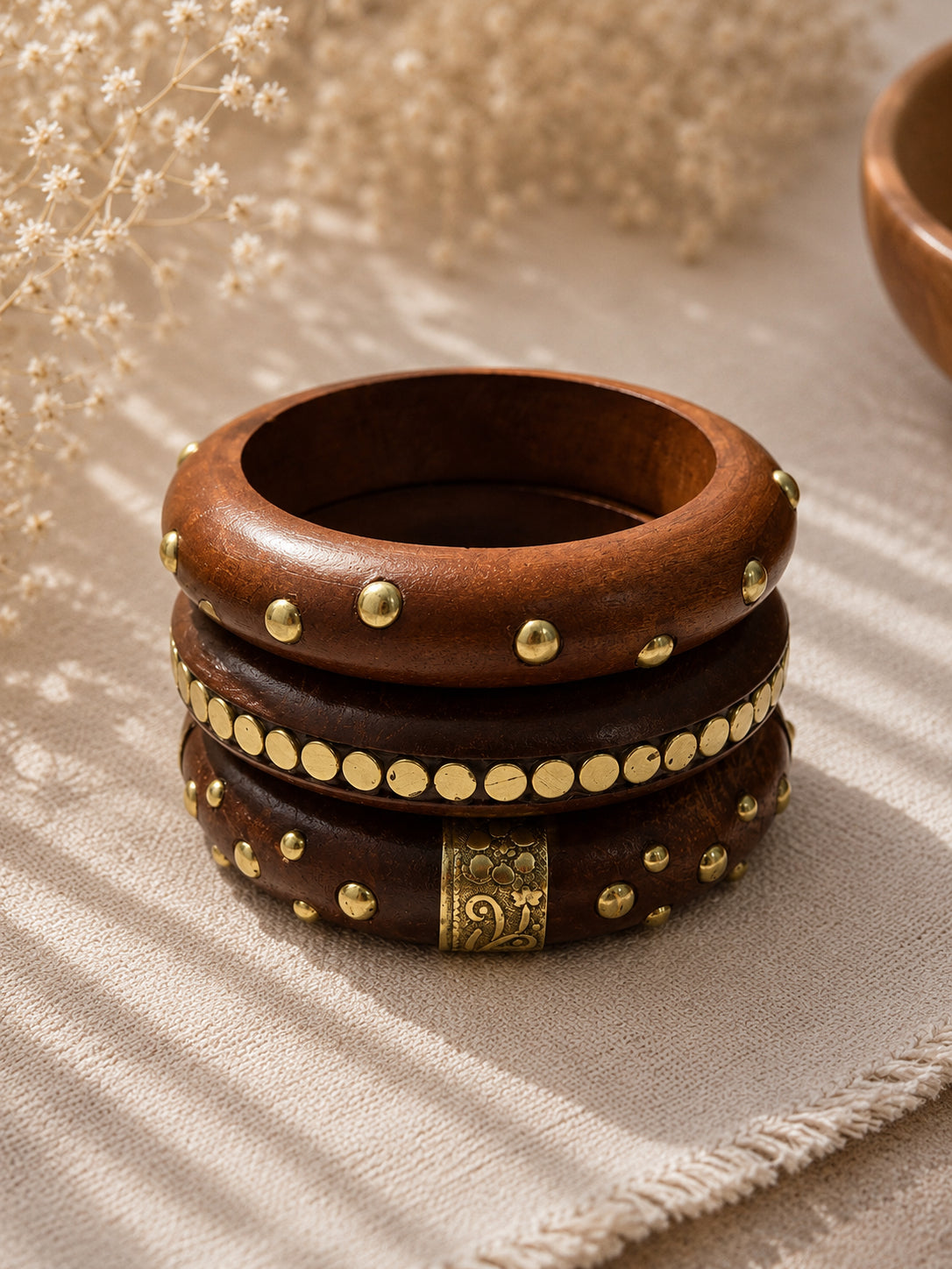 Stack of three brass-embellished wooden bangles on a linen cloth with dried white flowers in the background