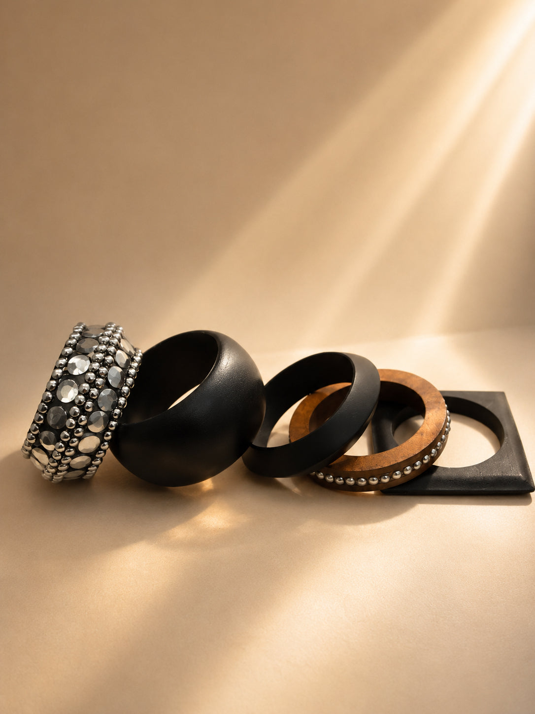Five wooden bangles laid flat on a beige surface including a wide oxidised silver embellished cuff and a square-edged black bangle