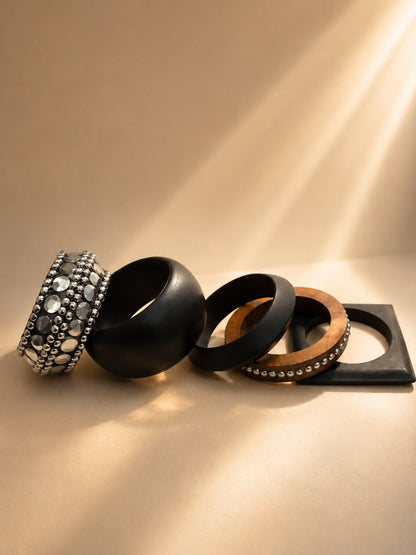 Five wooden bangles laid flat on a beige surface including a wide oxidised silver embellished cuff and a square-edged black bangle