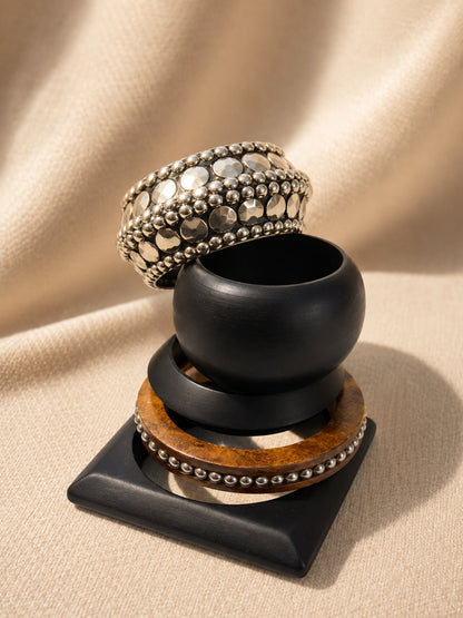 Stacked oxidised silver embellished cuff atop chunky black wooden bangle and walnut beaded bangle on a black square display stand against draped fabric
