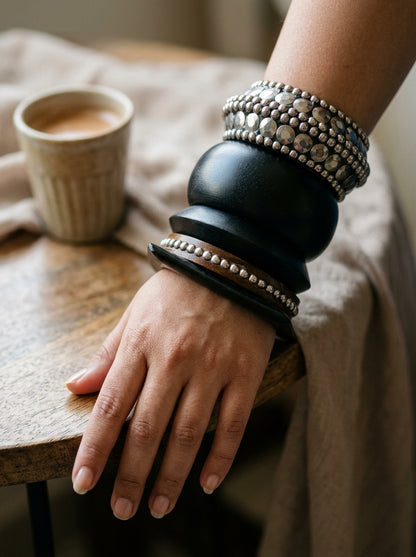 Close-up of a woman's wrist wearing a full stack of wooden and oxidised silver bangles resting on a rustic wooden table beside a coffee cup