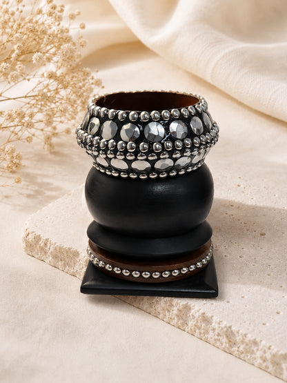 Front view of wide oxidised silver stone-embellished bangle stacked over black wooden bangle and walnut beaded bangle on a stone surface with dried flowers