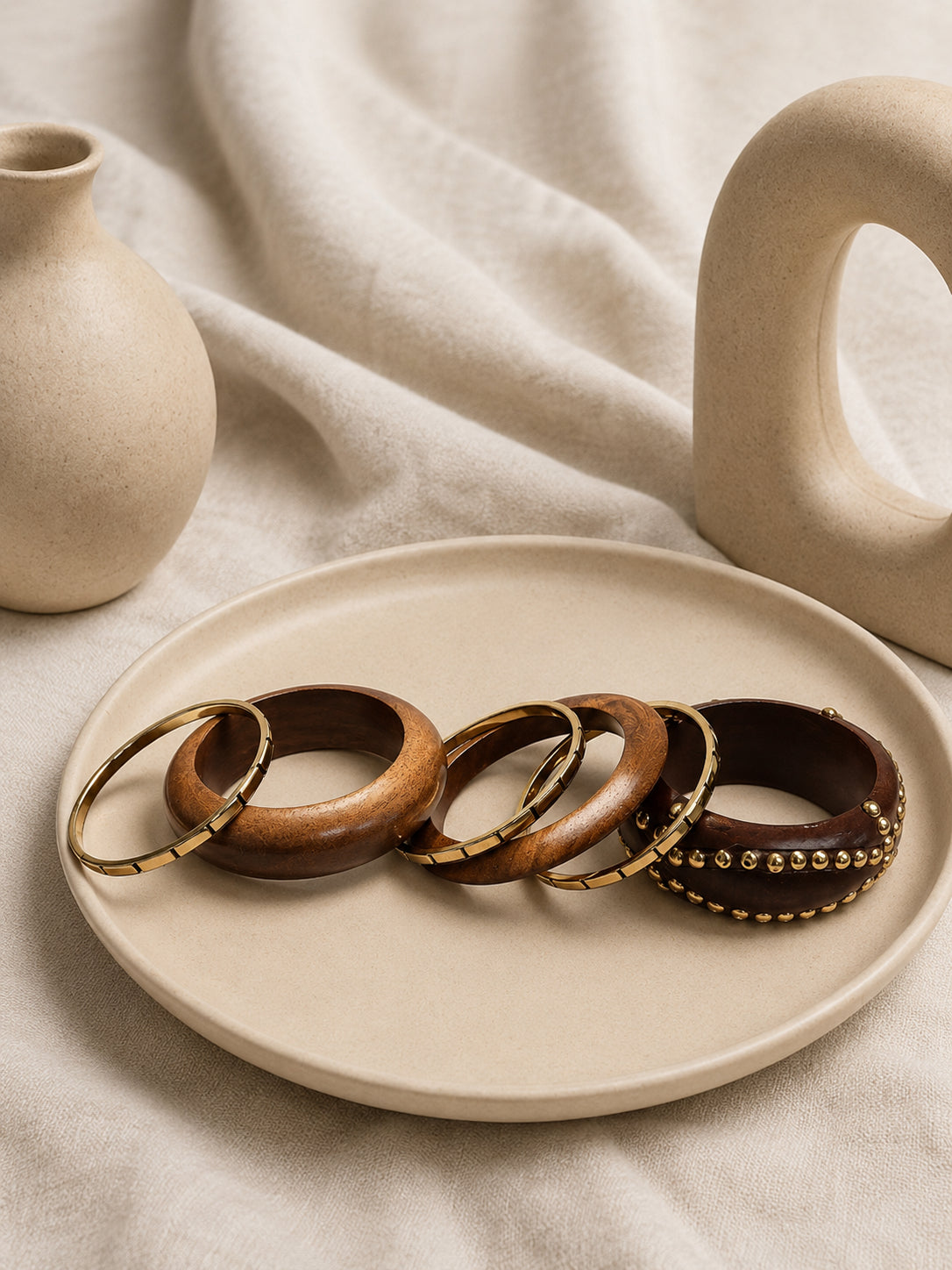 Three brown wooden bangles with gold-tone inlay displayed on a cream ceramic plate beside minimalist ceramic vases