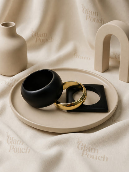 Three black wood and gold-tone bangles arranged on a beige ceramic tray against a branded cream fabric backdrop