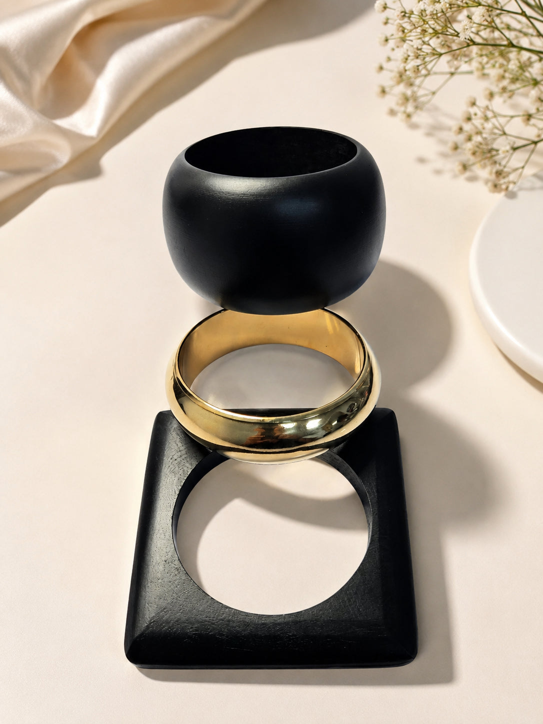 Black dome bangle, gold-tone round bangle and square black cuff stacked upright on a beige surface with baby's breath flowers