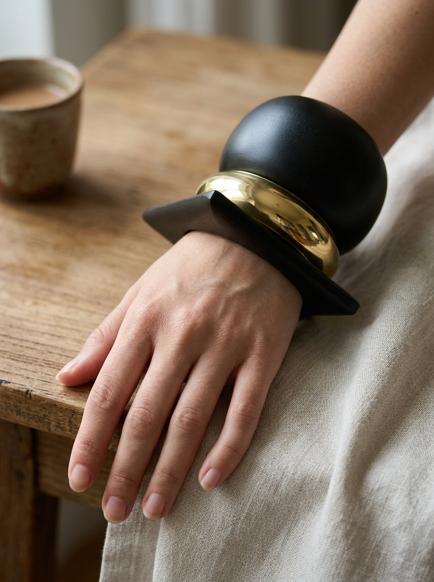 Woman wearing all three black and gold-tone geometric bangles stacked on wrist resting on a wooden table