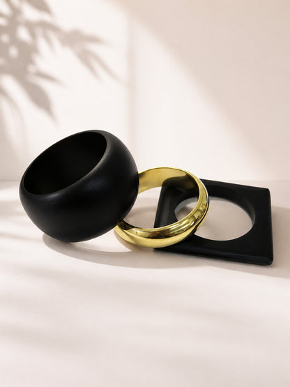 Black dome bangle leaning against gold-tone and square black cuff bangles on a soft beige surface with leaf shadow