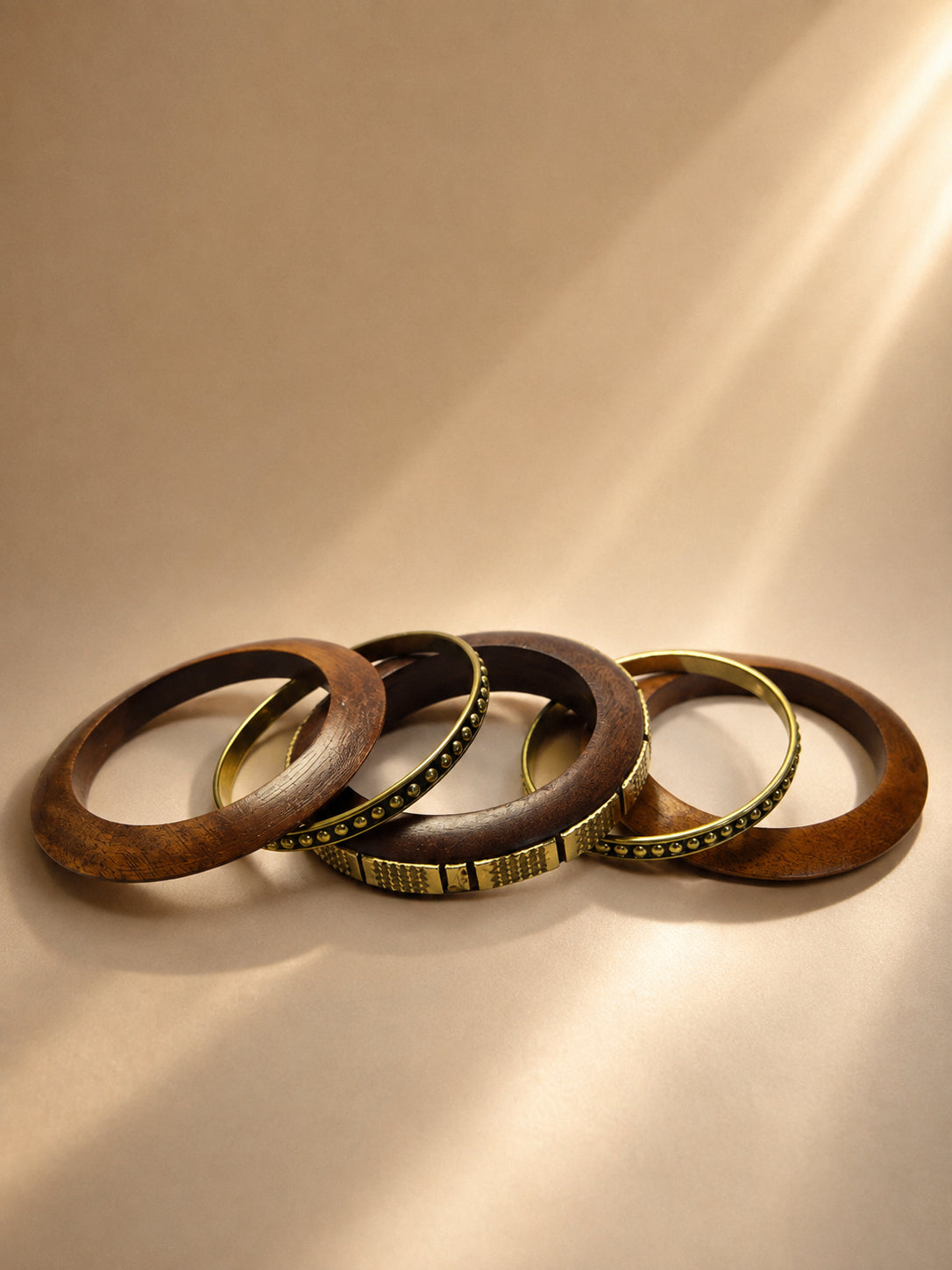 Five wooden and antique brass bangles fanned out on a warm beige surface in soft directional light
